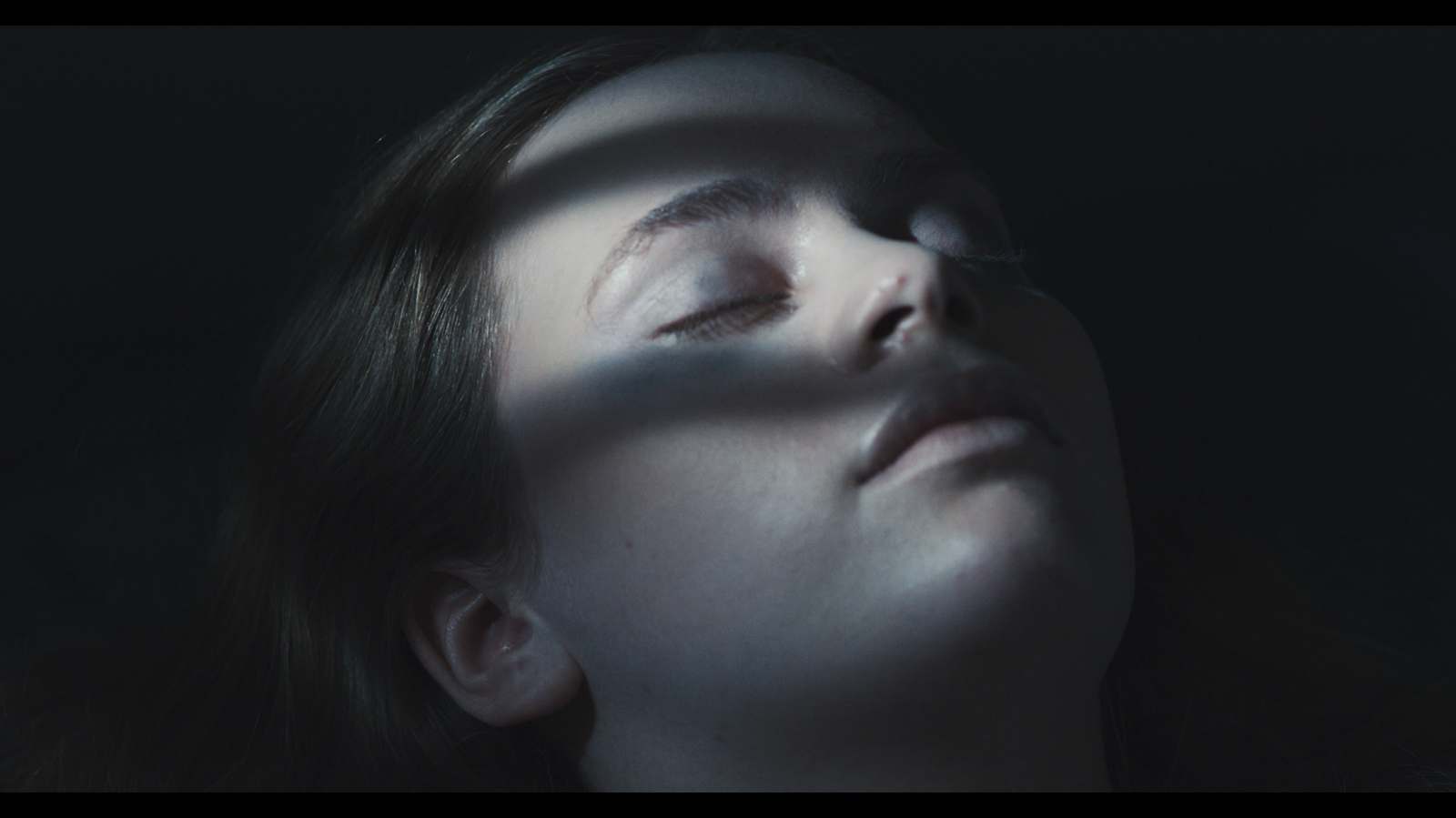 A face lit by narrow bands of light in a still from After the Body