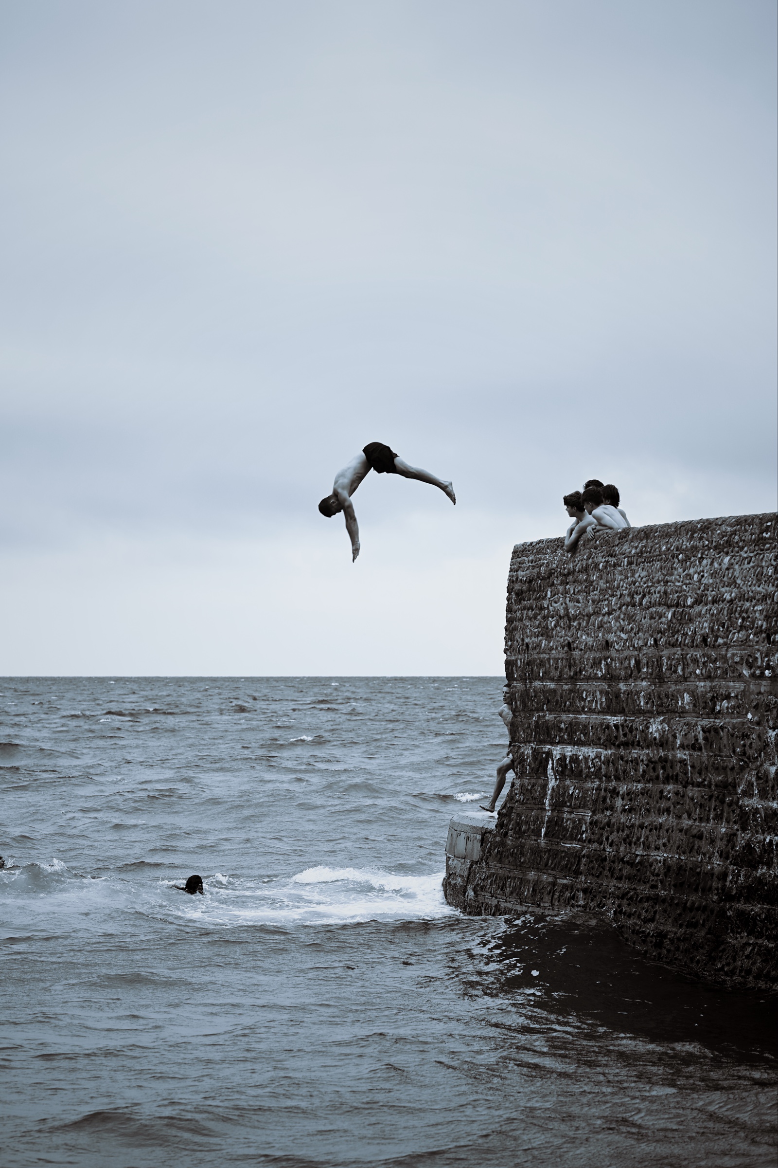 A diver arches backwards above the water beside the stone wall