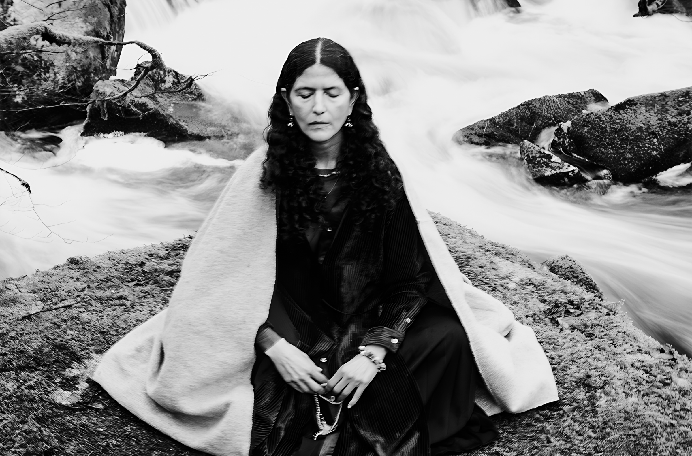 A seated figure wrapped in pale cloth beside flowing water in black and white